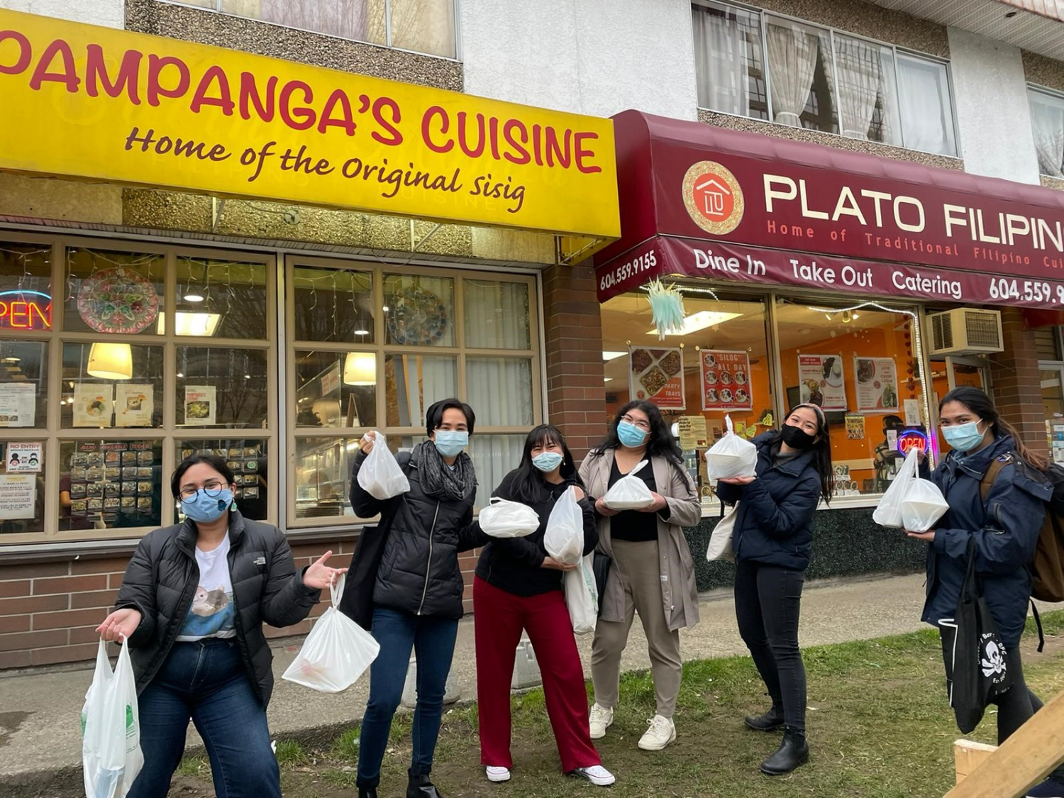 A group of smiling masked people holding bagged takeout containers standing in front of two Philippino restaurants